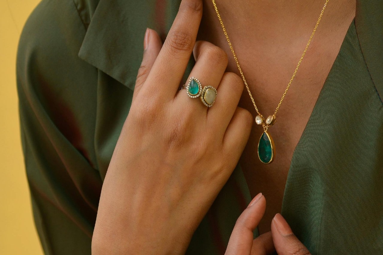 A woman wearing a two gemstone ring with halos around each center stone that matches the gemstone and diamond pendant necklace she’s wearing