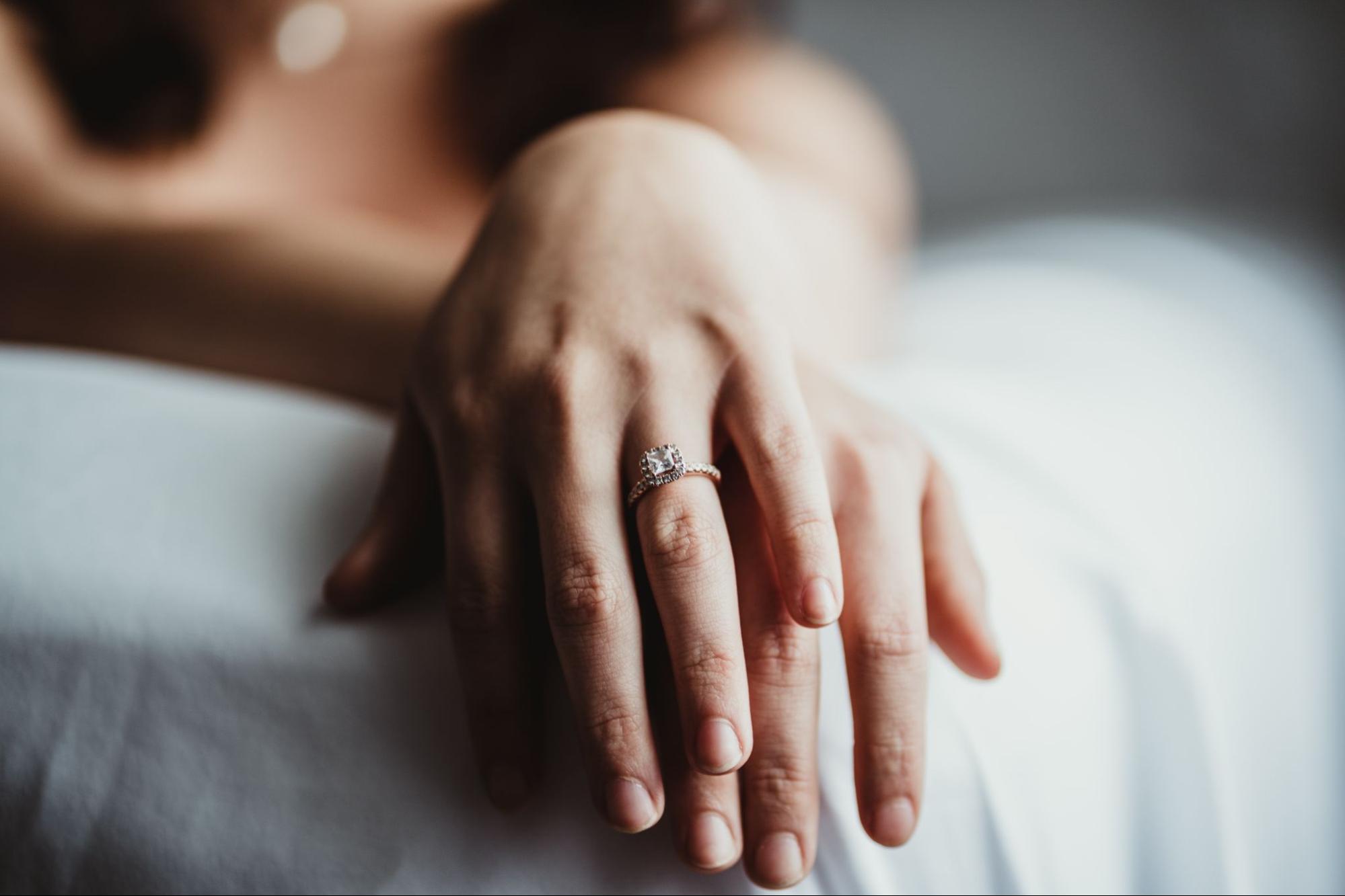 A close up of two hands stacked on top of each other while laying on a bed, showcasing a beautiful engagement ring.