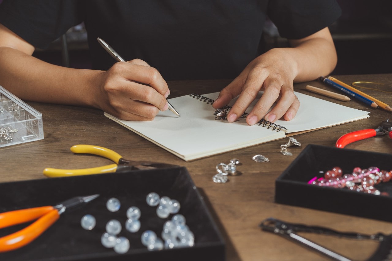 A person wearing a black shirt sitting at a desk and writing on a blank notebook with jewelry tools spread across the table.