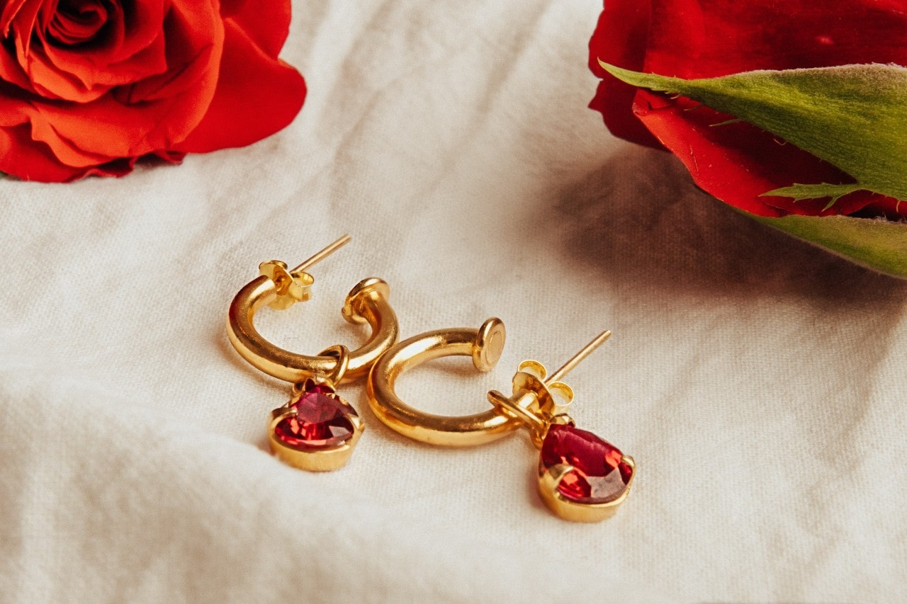 a pair yellow gold huggie/drop earrings featuring red gemstones on a piece of cream linen