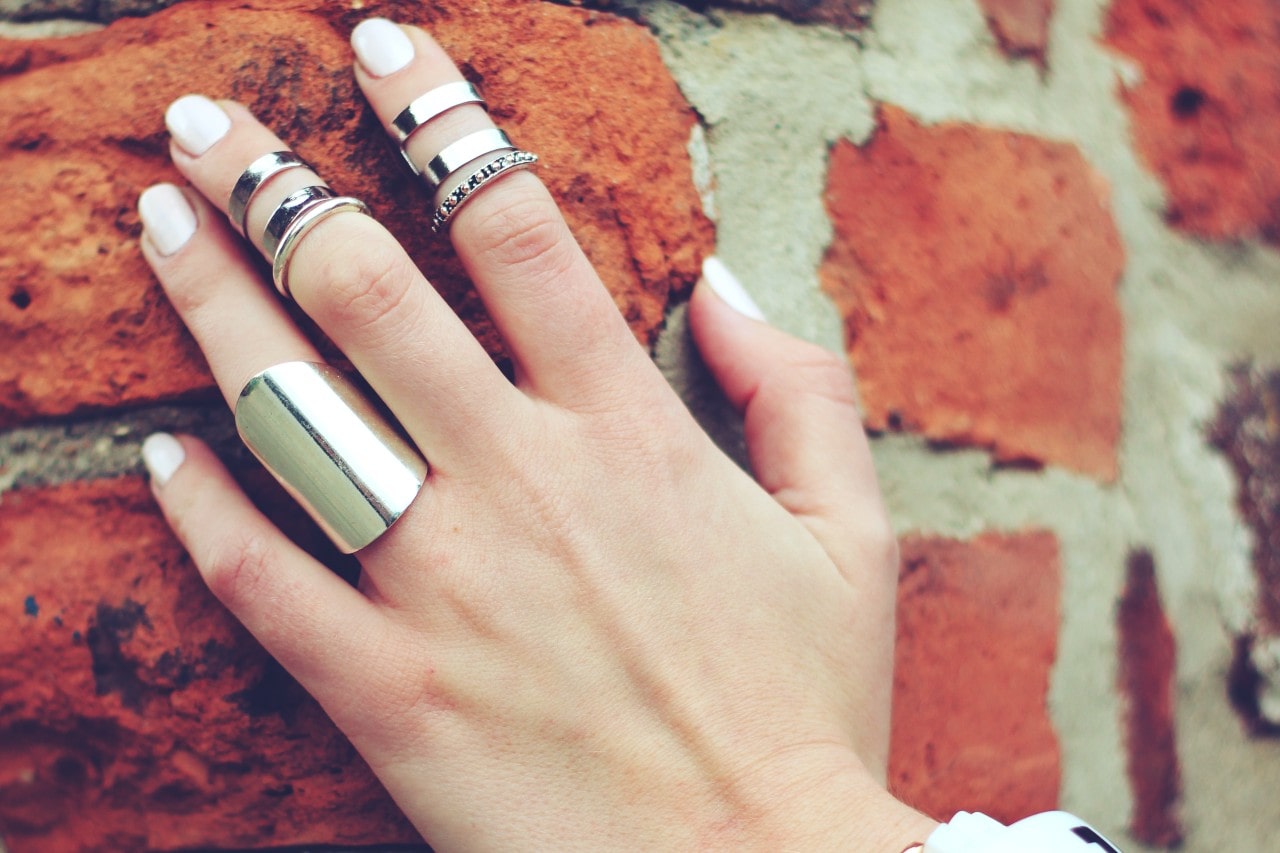 Close-up of a hand with white nails against a red brick wall, wearing multiple silver rings.