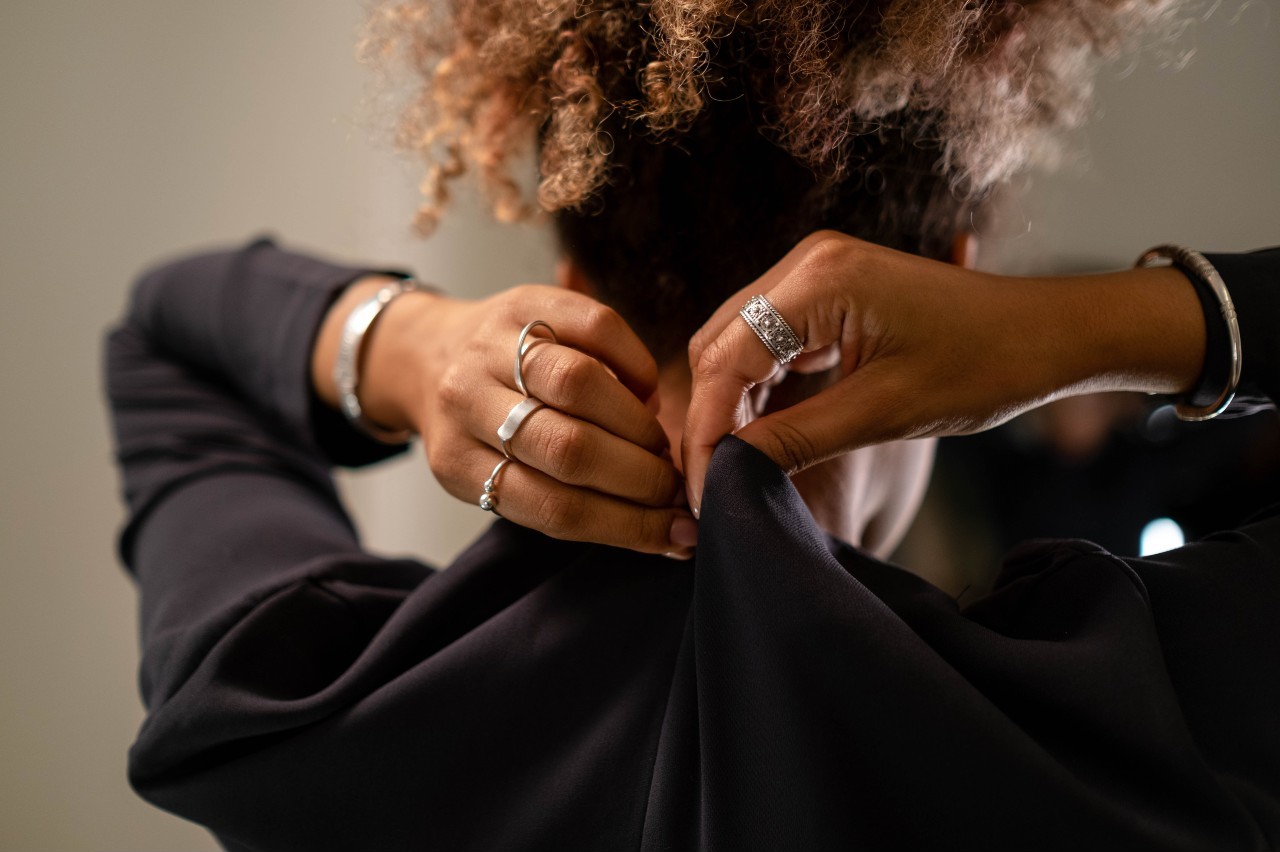 A person with curly hair in a black long-sleeve shirt wearing multiple silver rings and bracelets adjusts the back of their shirt.