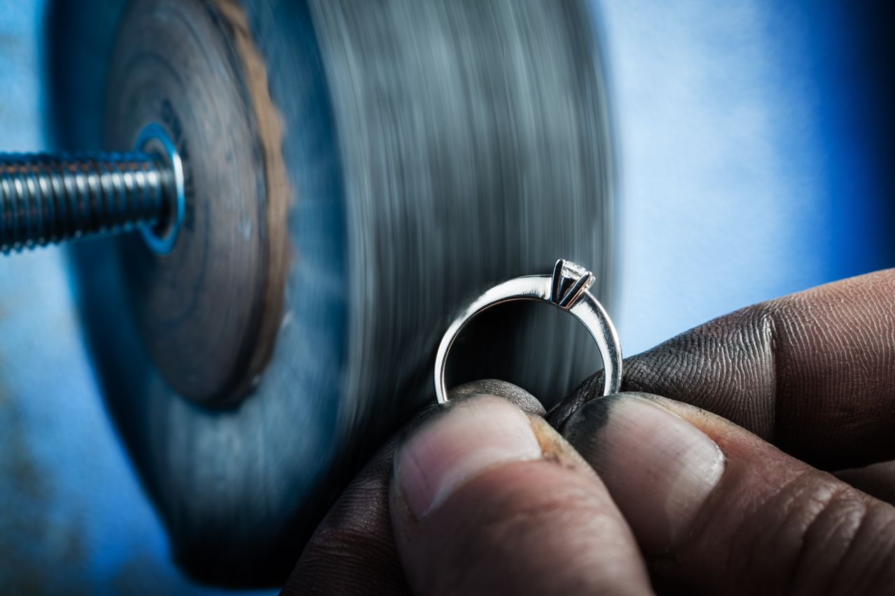 Close-up of a jeweler’s hand polishing a silver ring on a spinning wheel against a blue-tinted background.