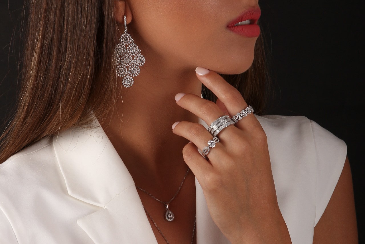 A chic woman in a white top wearing diamond earrings, a necklace, and rings against a dark background.