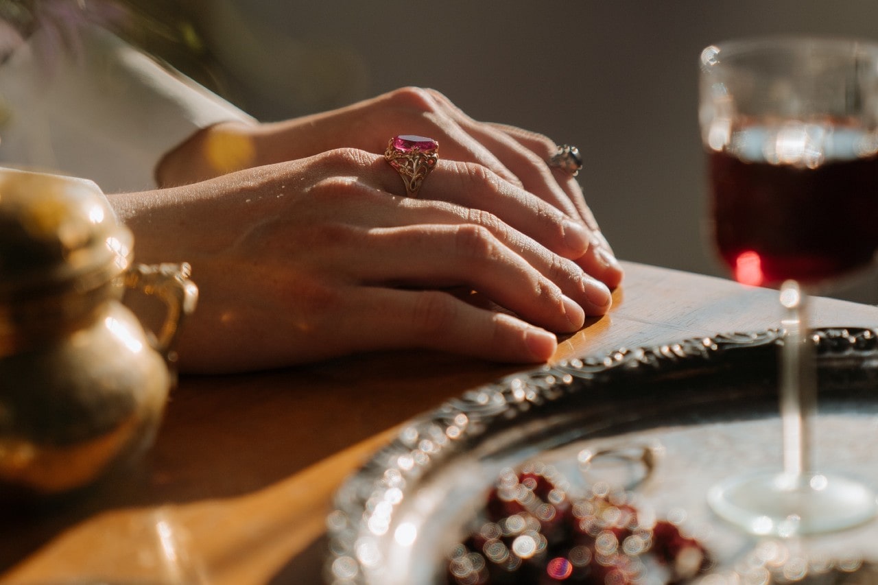 a pair of hands resting on a table, wearing gemstone fashion rings, next to a silver platter and glass of wine