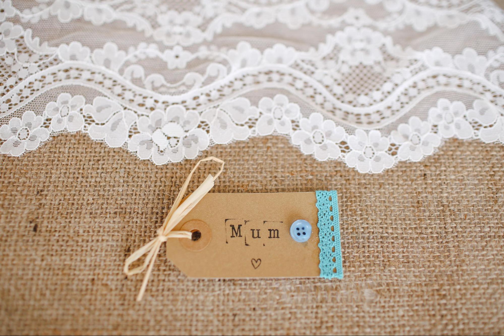 A gift tag for a mum sat on top of a rough woven cloth and some white lace.