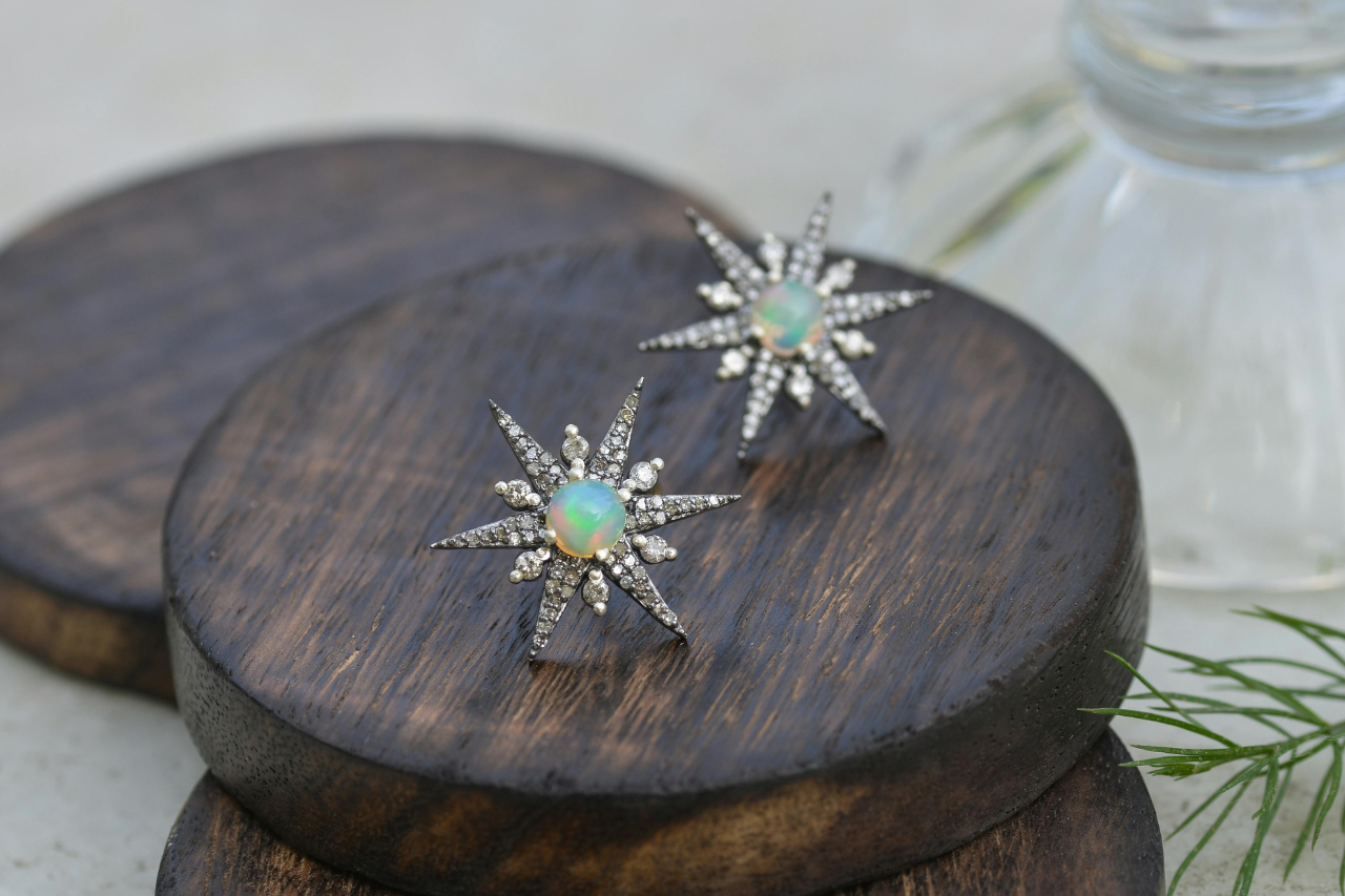 A close-up of gorgeous starburst-shaped opal and diamond stud earrings displayed on stacked dark wood discs.