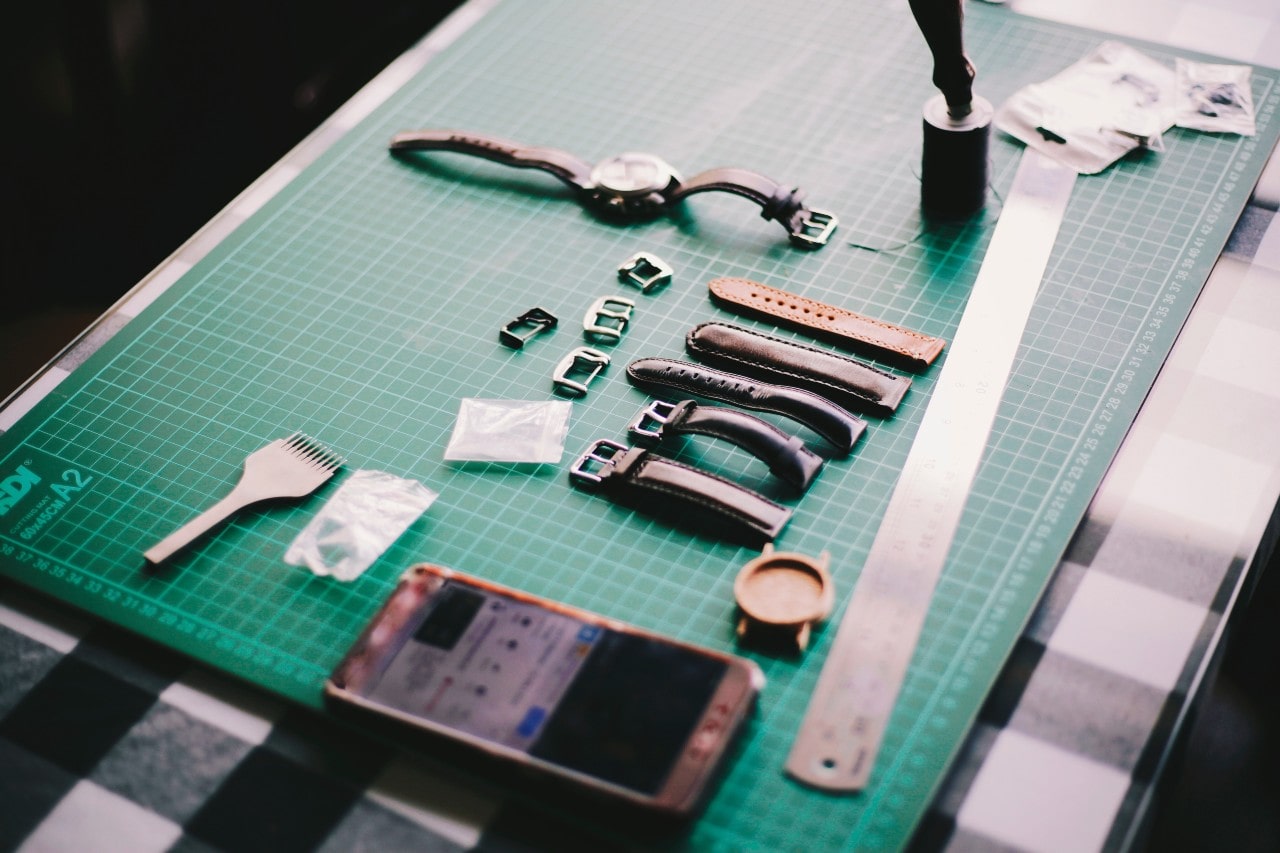 Several leather watch straps are placed on a green table near a smartphone and watch.