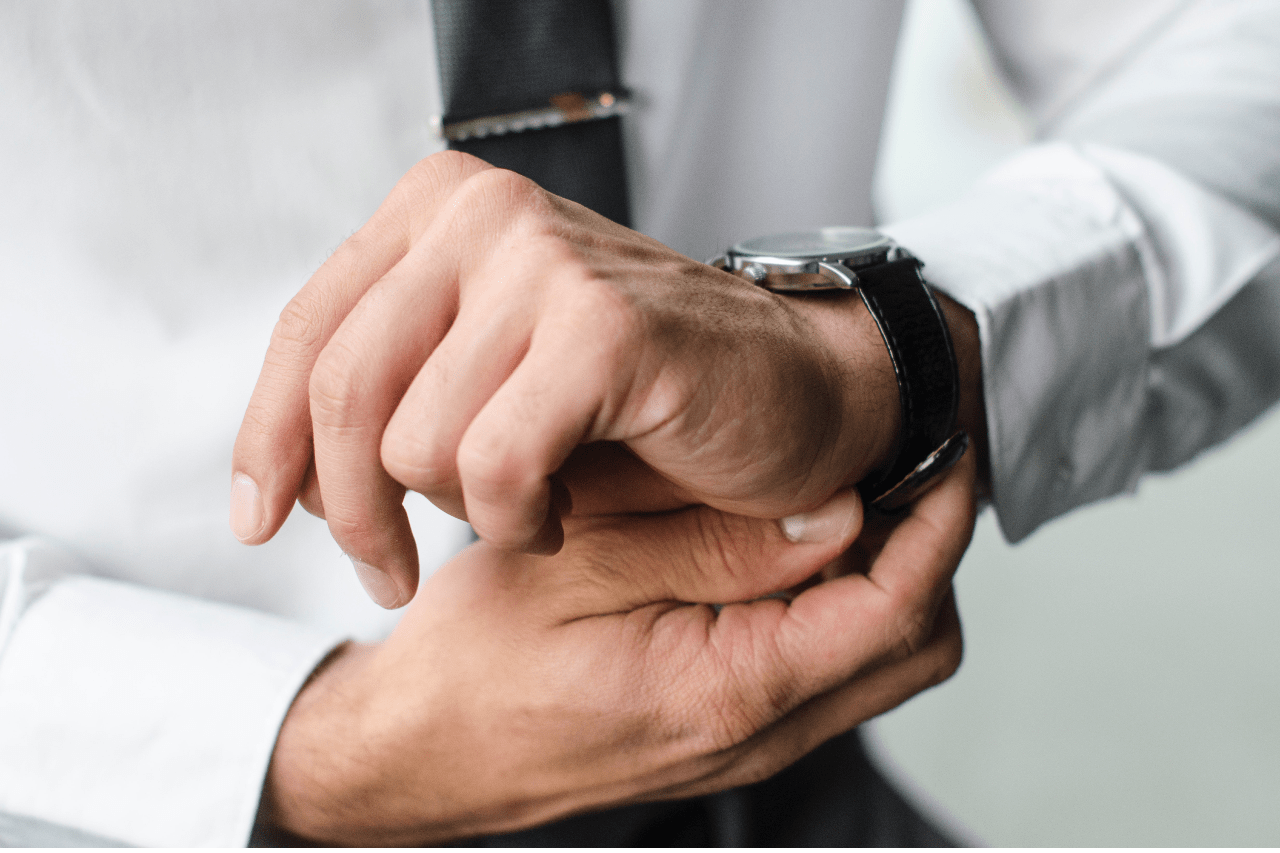 A close-up of a man's hands in a white shirt adjusting a black leather-strap wristwatch