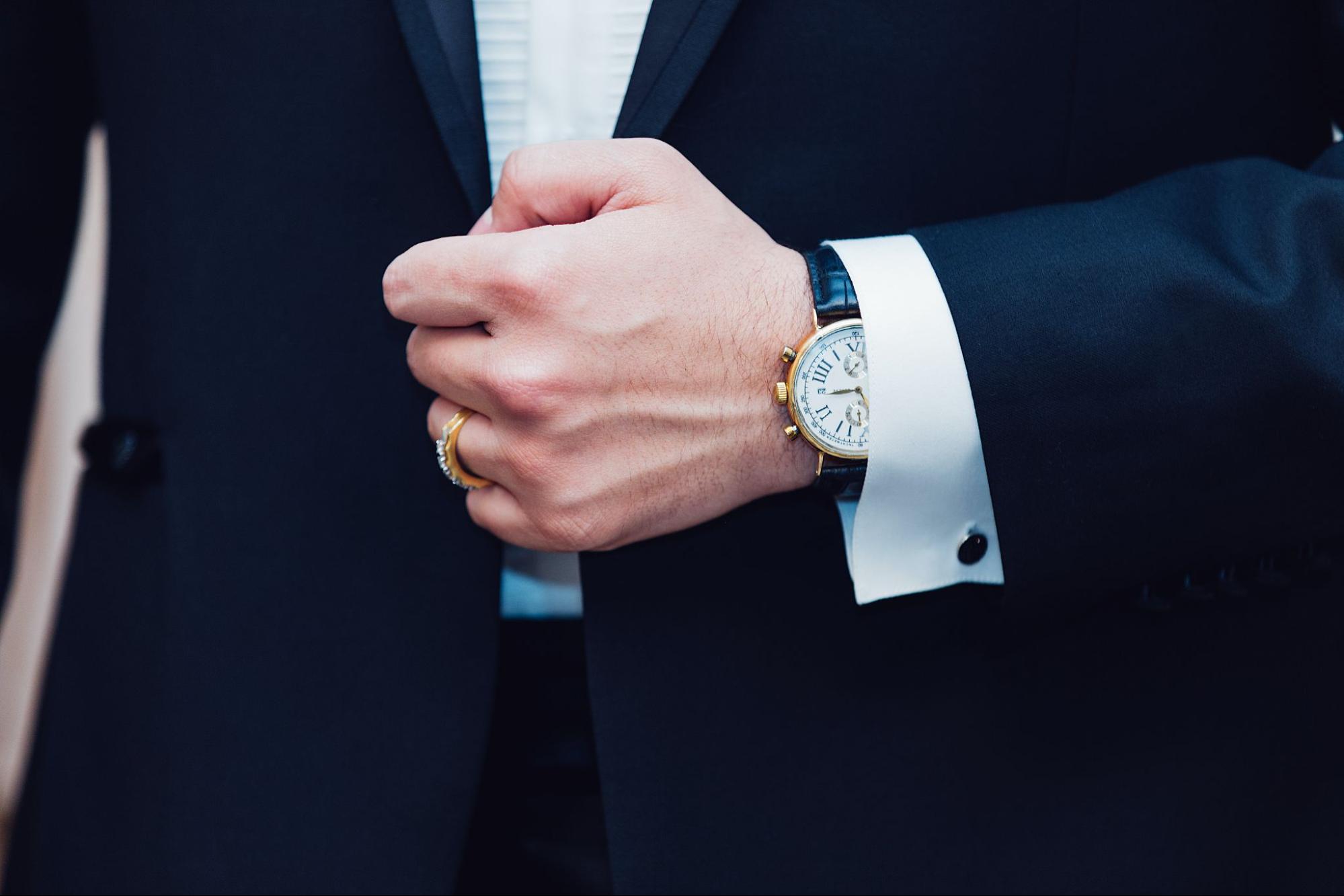A close up of a man’s hand in navy suit, showcasing a watch with white dial and black leather strap and yellow gold wedding band.