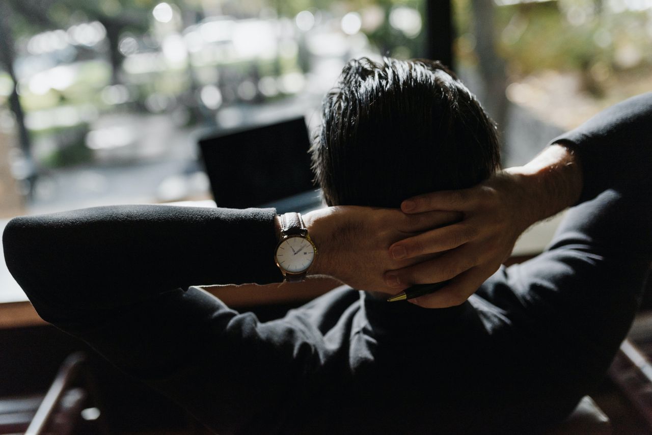 A man in a dark sweater relaxes with his hands behind his head, showcasing a classic wristwatch, while sitting in front of a computer with a blurry outdoor view.