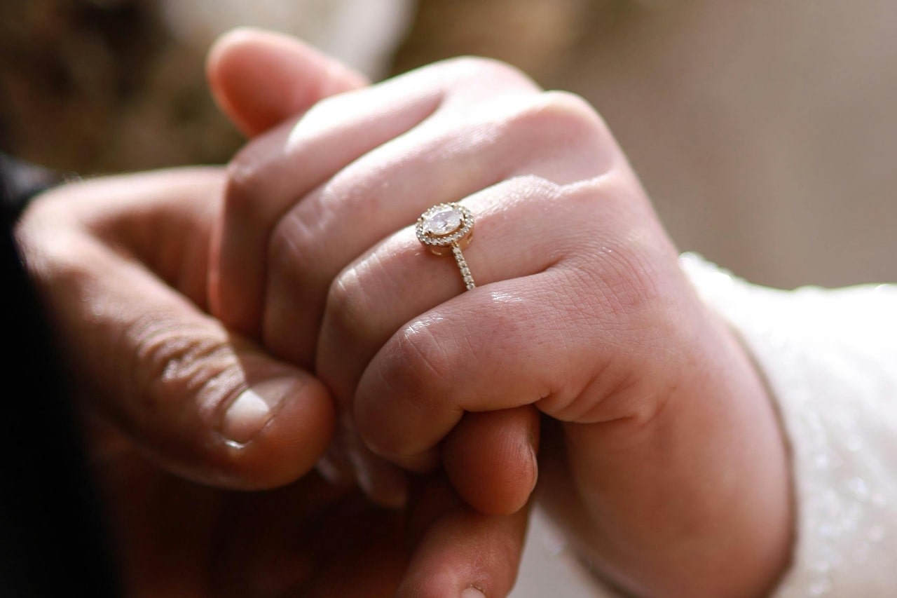A close-up of two hands clasped together, highlighting a halo-style diamond engagement ring with a pavé band on one finger.