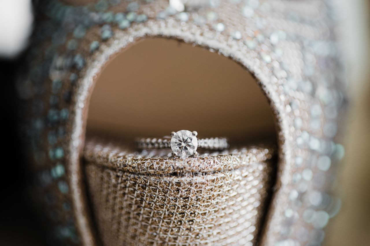 A sparkling diamond ring rests elegantly on a glittering, rhinestone-adorned high heel shoe.