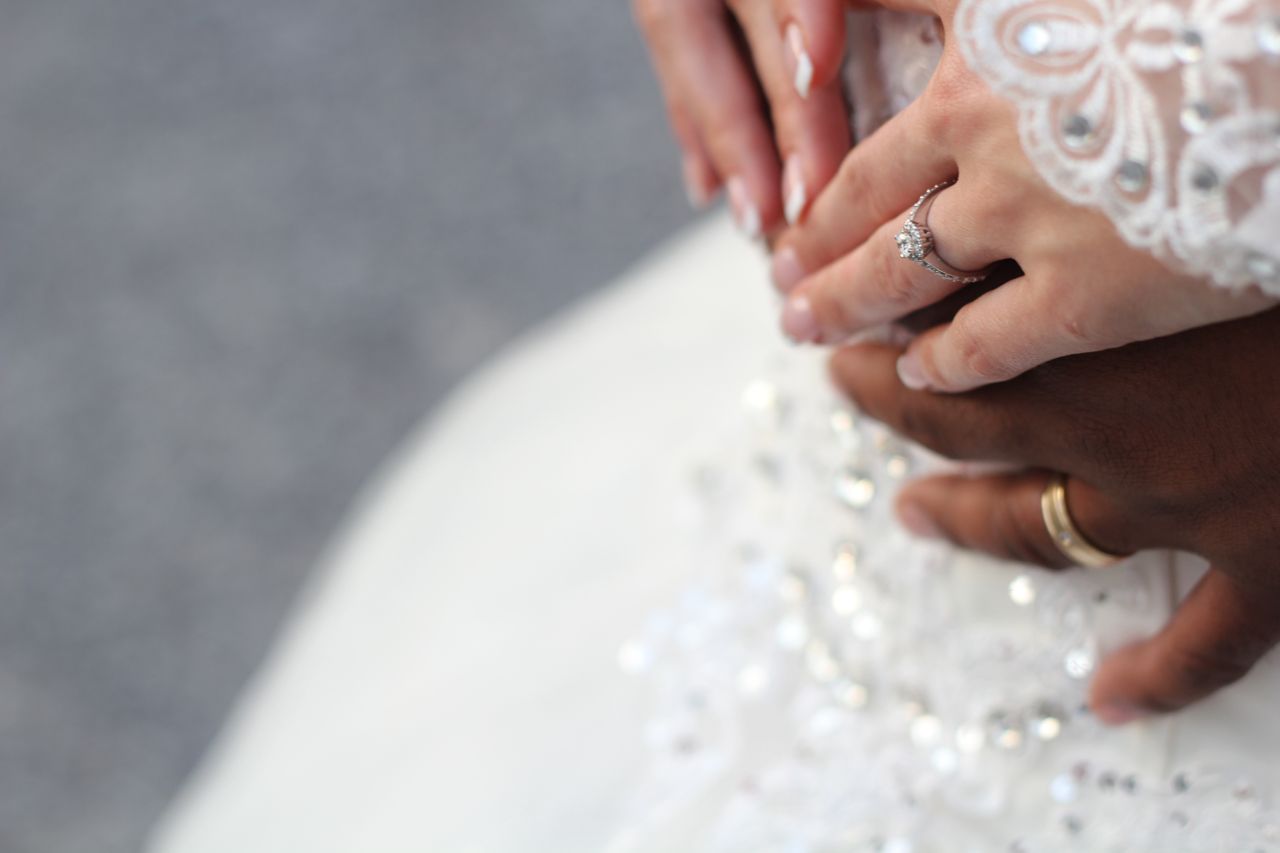 Close-up of a bride and groom’s hands resting on a white beaded dress, the bride’s hand adorned with a sparkling ring and the groom wearing a gold band.