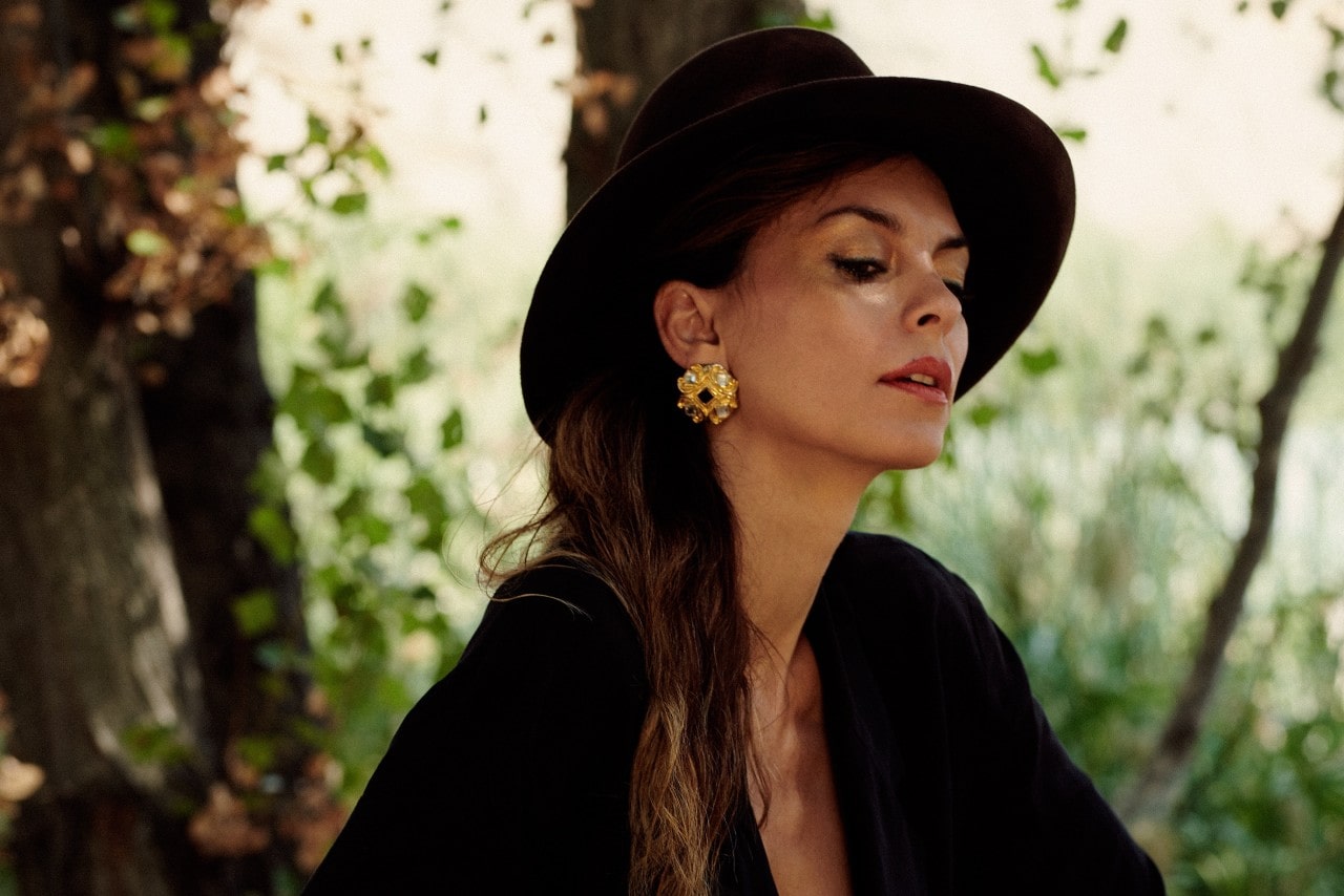 A close-up of a woman in a dark hat and bold gold earrings in front of a leafy tree.