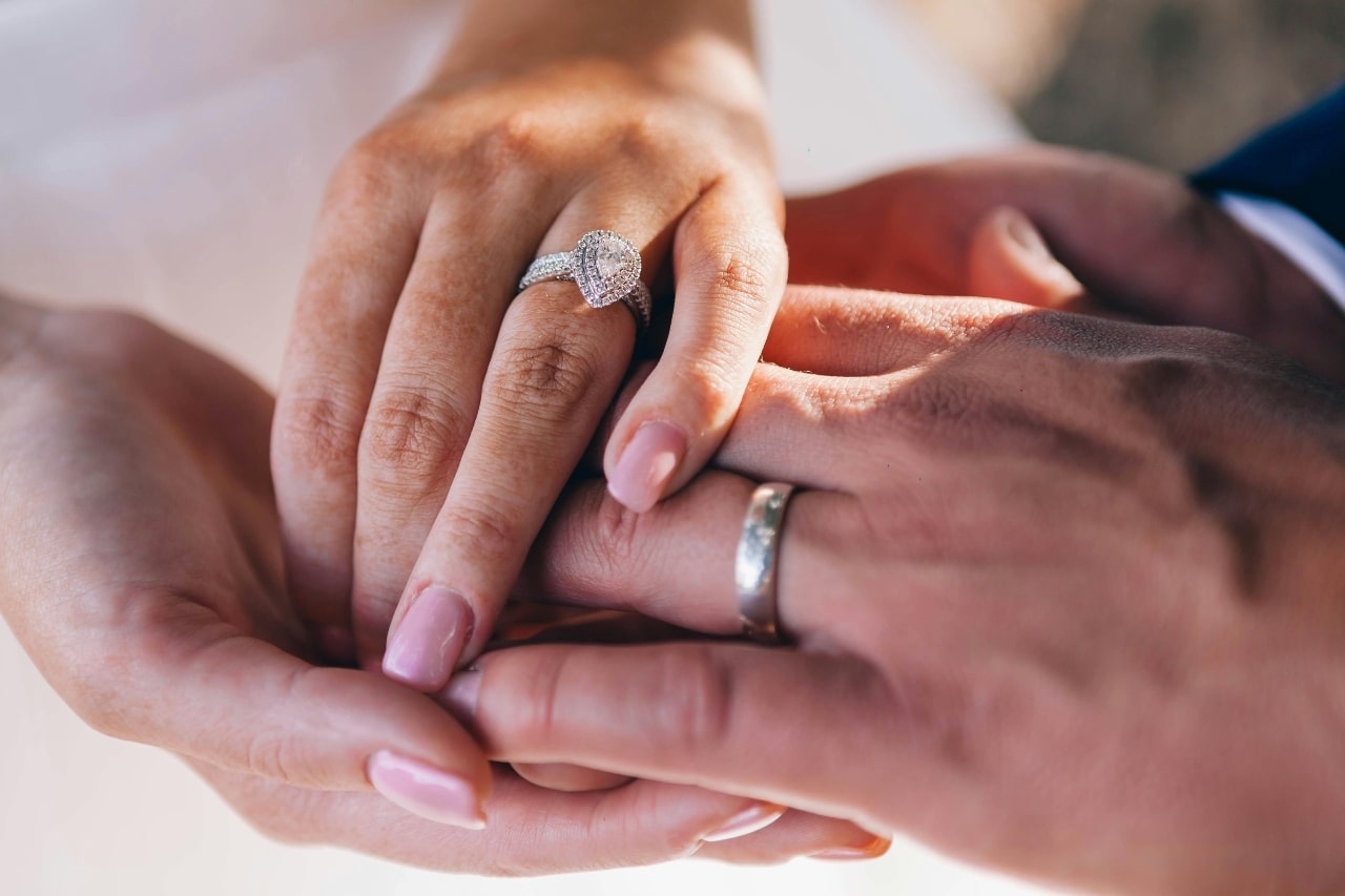 Close-up of a woman's hand with a diamond engagement ring gently held by a man's hand wearing a wedding band.