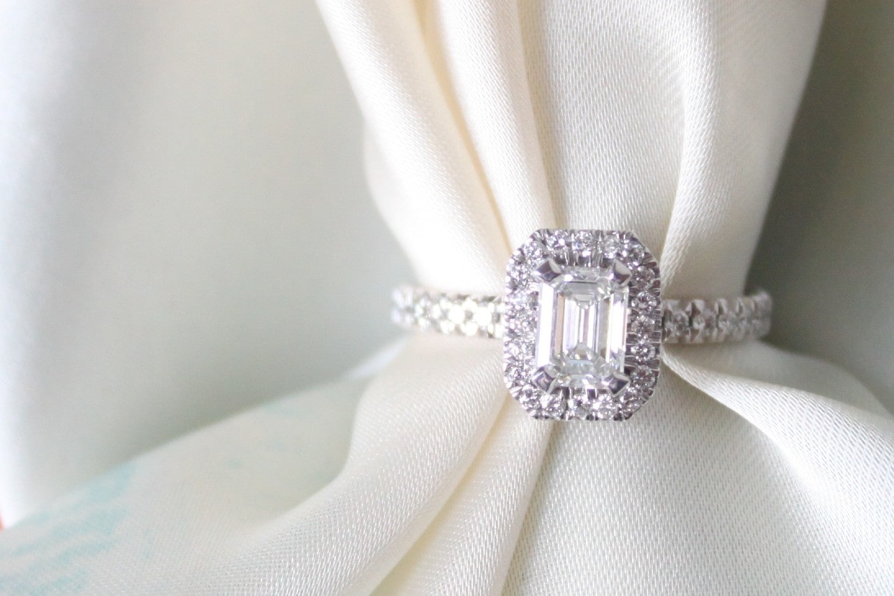 A sparkling emerald-cut diamond ring with halo setting rests elegantly on a soft, white fabric.
