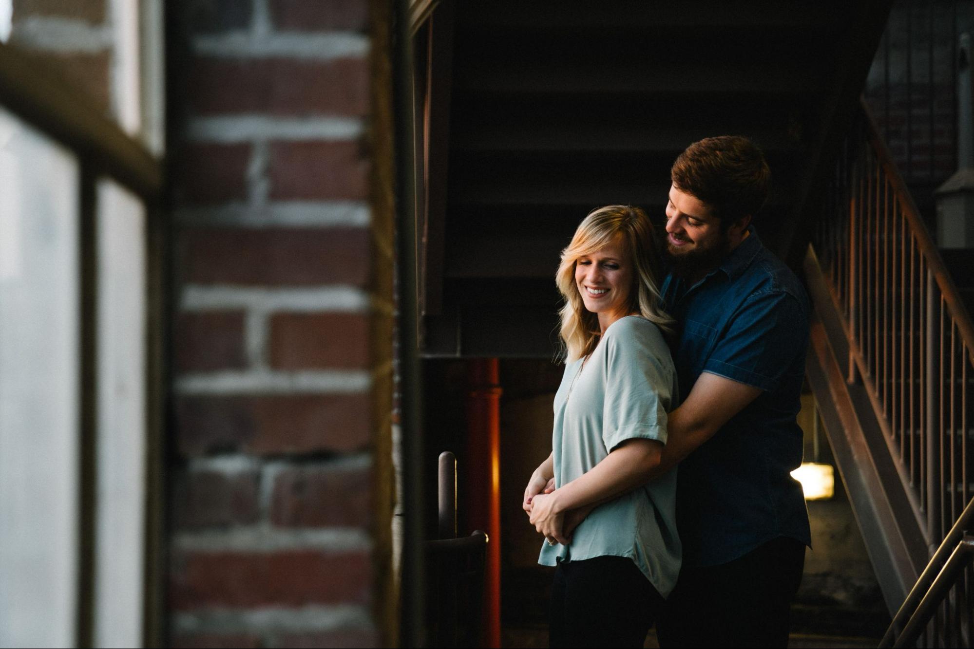 A couple embraces under the stairs in a brick building in front of the window.