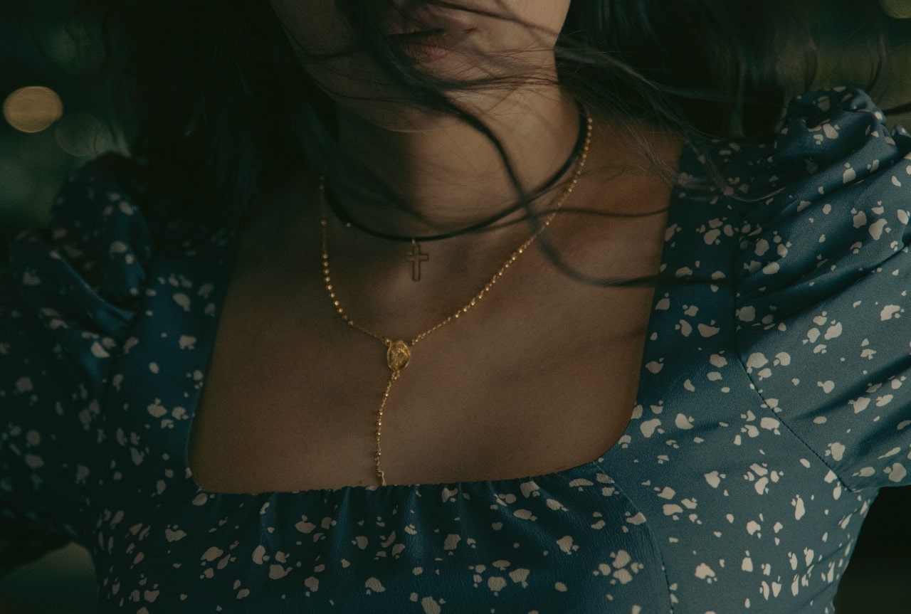A woman wearing a blue patterned dress is shown with her hair blowing softly as she displays yellow gold lariat and black cross pendant.