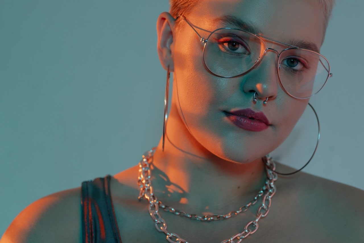 A person with short hair, wearing large glasses, hoop earrings, and a chain necklace, gazes into the camera under soft blue and orange lighting.