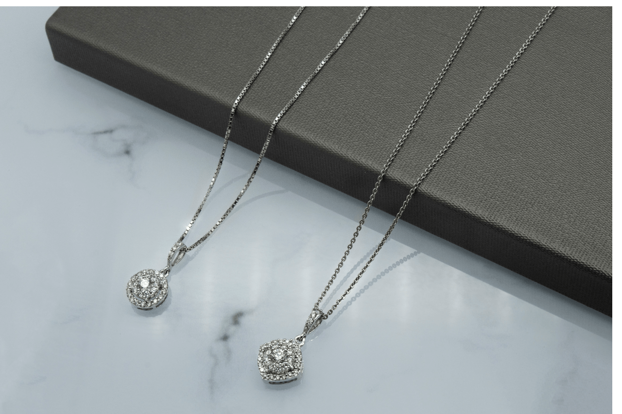 Two silver necklaces with diamond pendants resting on a gray box, placed against a marble background.