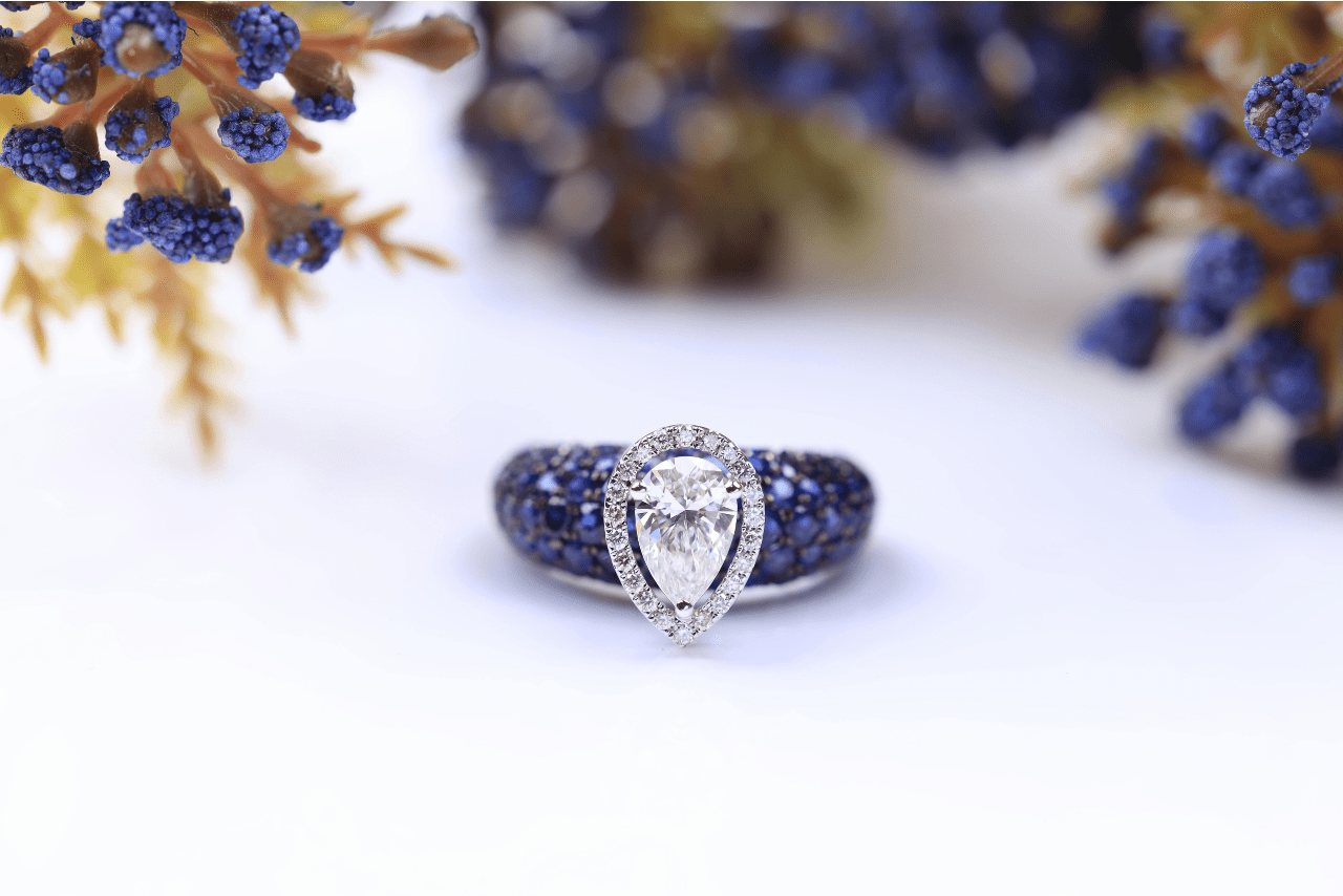 A pear shape engagement ring with a diamond halo and a dark blue side stone band, blue florals in the background.