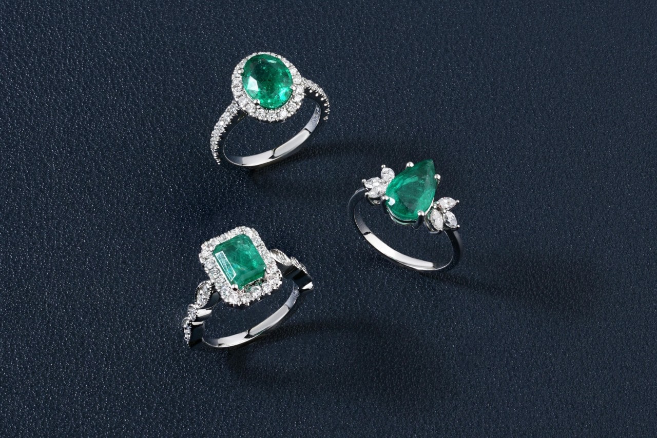 Three emerald engagement rings, one a round stone, one an emerald, and one a pear, standing up on a navy leather surface.
