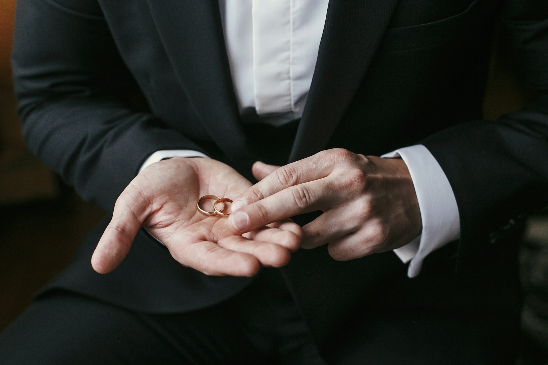 A close up of two thin gold wedding bands resting on a palm of the groom in a black suit with white shirt A close up of two thin gold wedding bands resting on a palm of the groom in a black suit with white shirt