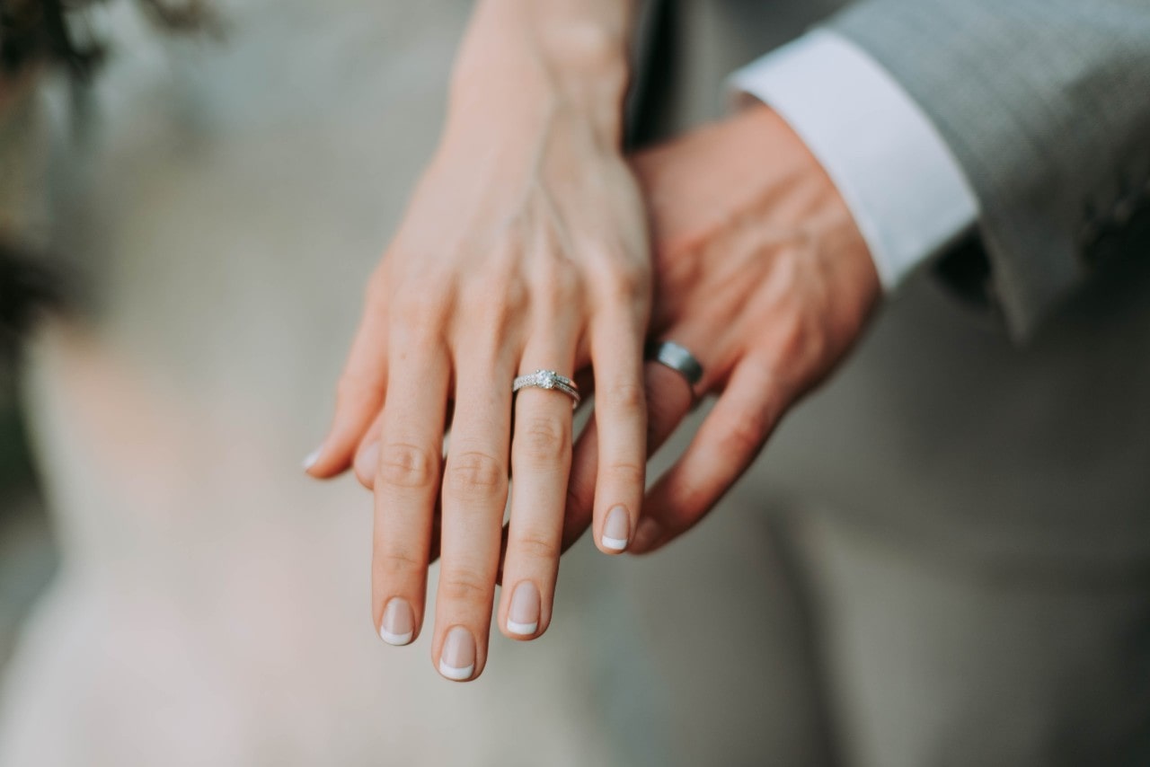 A photograph of a newlywed couple's hands showing their wedding rings, with the bride's French manicure and sparkling engagement ring over the groom's hand with silver wedding band A photograph of a newlywed couple's hands showing their wedding rings, with the bride's French manicure and sparkling engagement ring over the groom's hand with silver wedding band