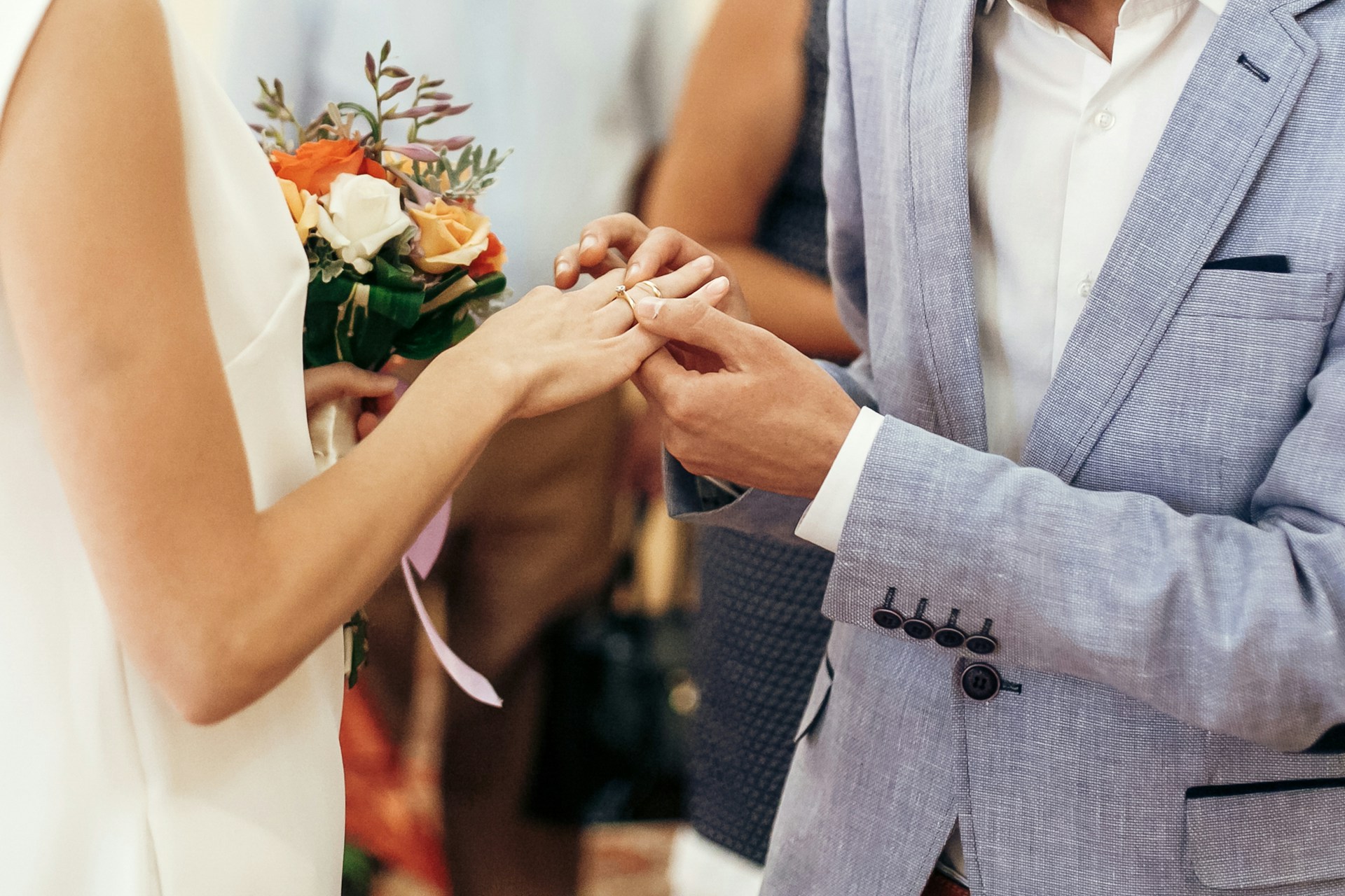 A groom in a light grey suit placing a gold wedding band on his bride's finger during a ceremony, with a bouquet of flowers in her other hand. A groom in a light grey suit placing a gold wedding band on his bride's finger during a ceremony, with a bouquet of flowers in her other hand.