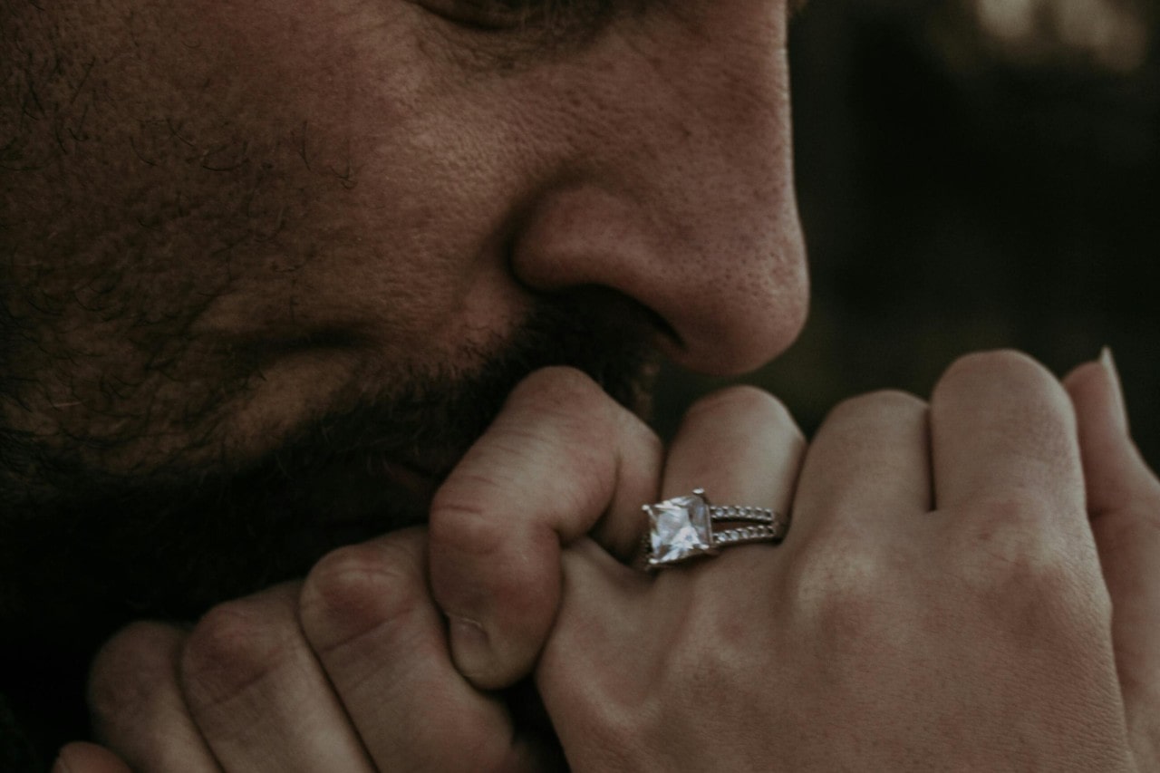 Close-up of a person gently kissing a hand adorned with an elegant square-cut diamond ring featuring a split band set with sparkling diamonds. Close-up of a person gently kissing a hand adorned with an elegant square-cut diamond ring featuring a split band set with sparkling diamonds.