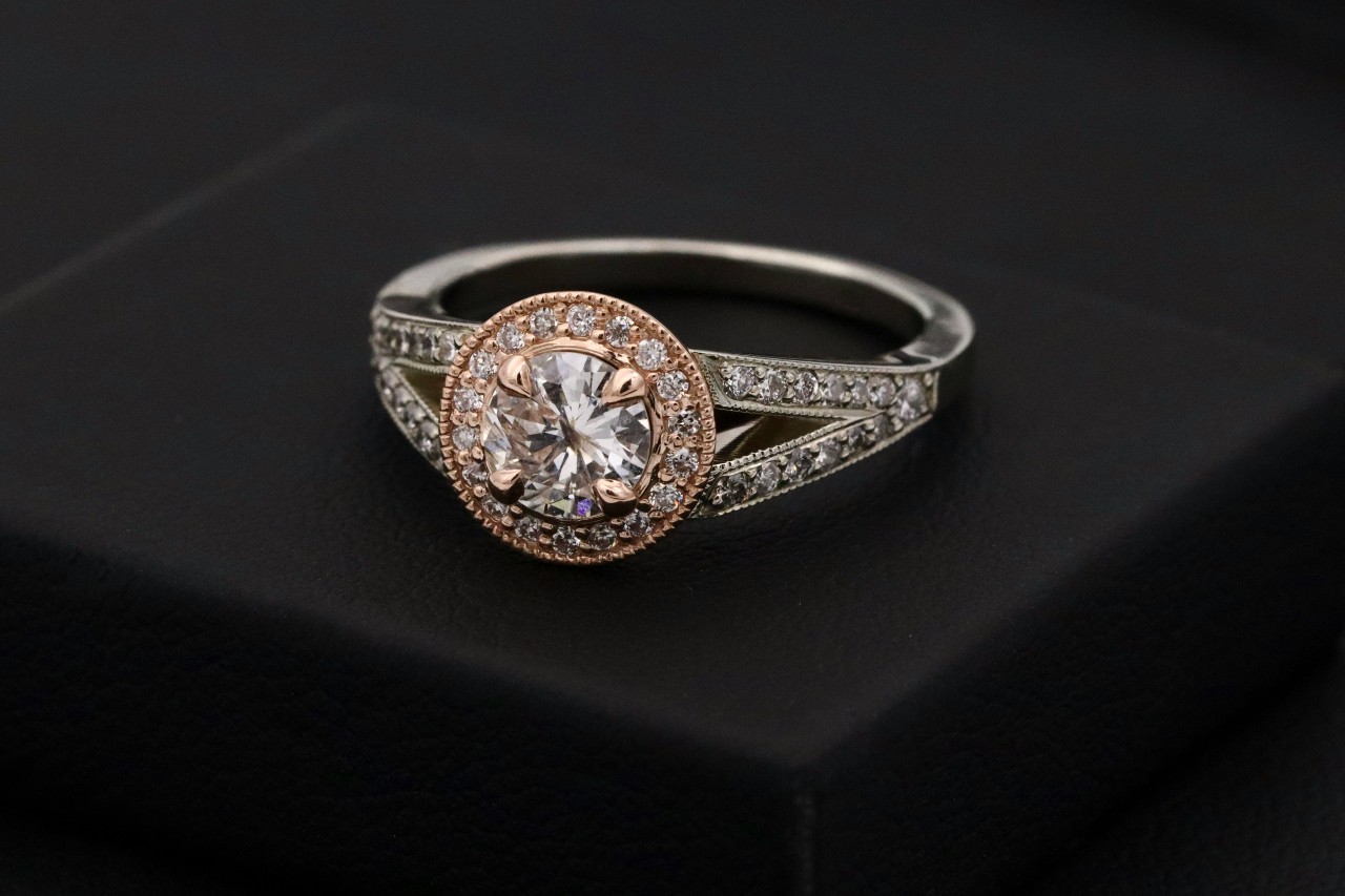 An white and rose gold ring featuring a central diamond encircled by a halo of smaller diamonds, with a split, diamond-adorned band resting on a black surface. An white and rose gold ring featuring a central diamond encircled by a halo of smaller diamonds, with a split, diamond-adorned band resting on a black surface.